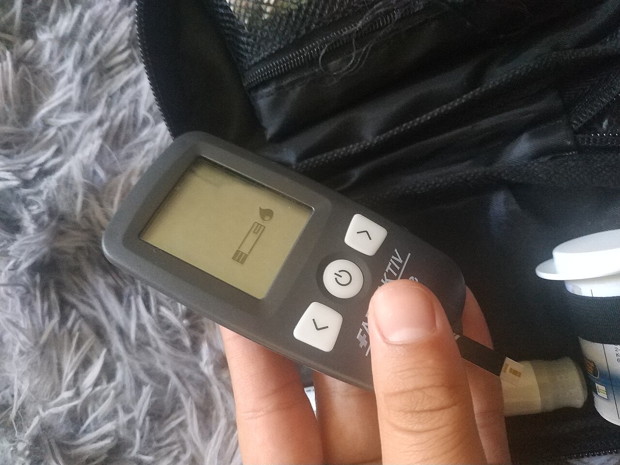 A hand holds a glucose meter showing a strip and drop symbol beside a strip container and lancing device.