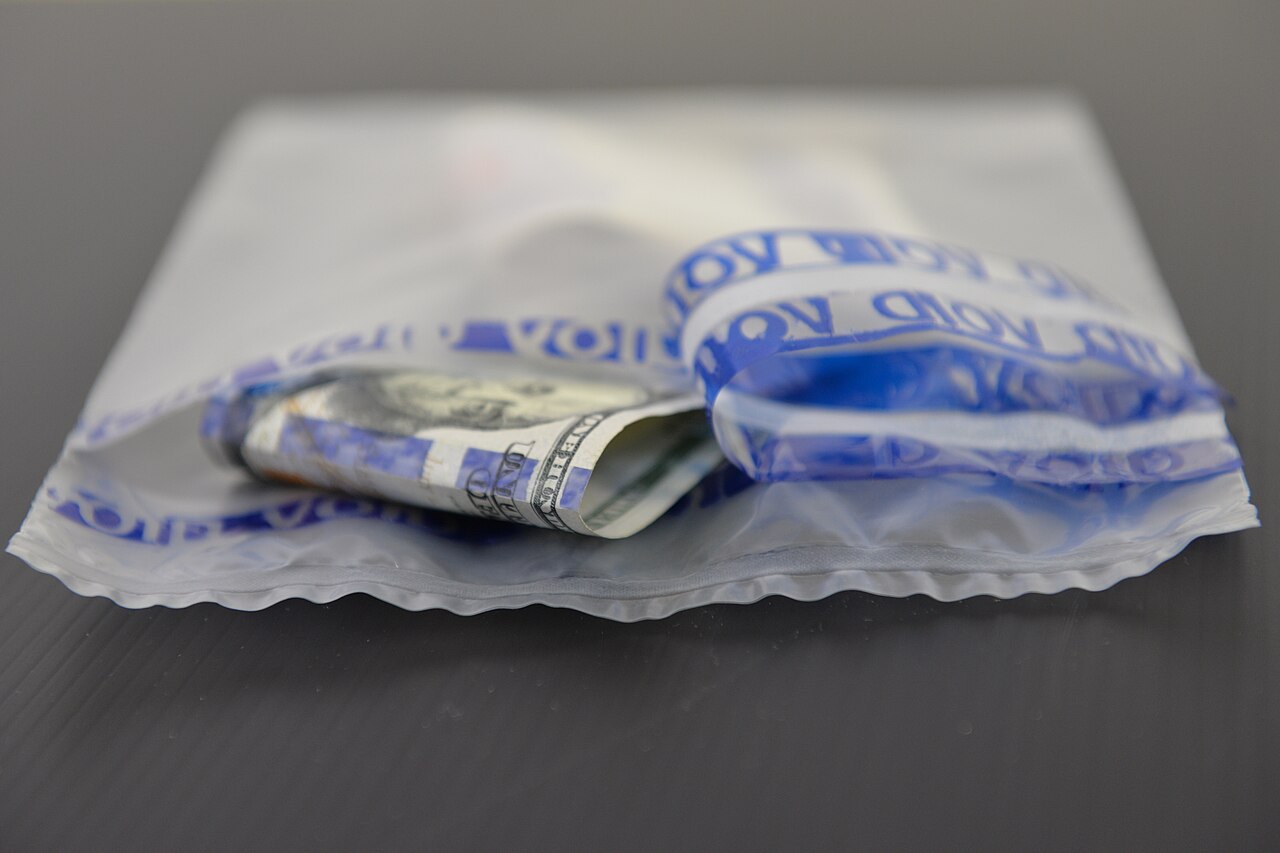 A clear security bag containing cash has a partly peeled blue seal with visible VOID lettering; no patient or specimen is shown.